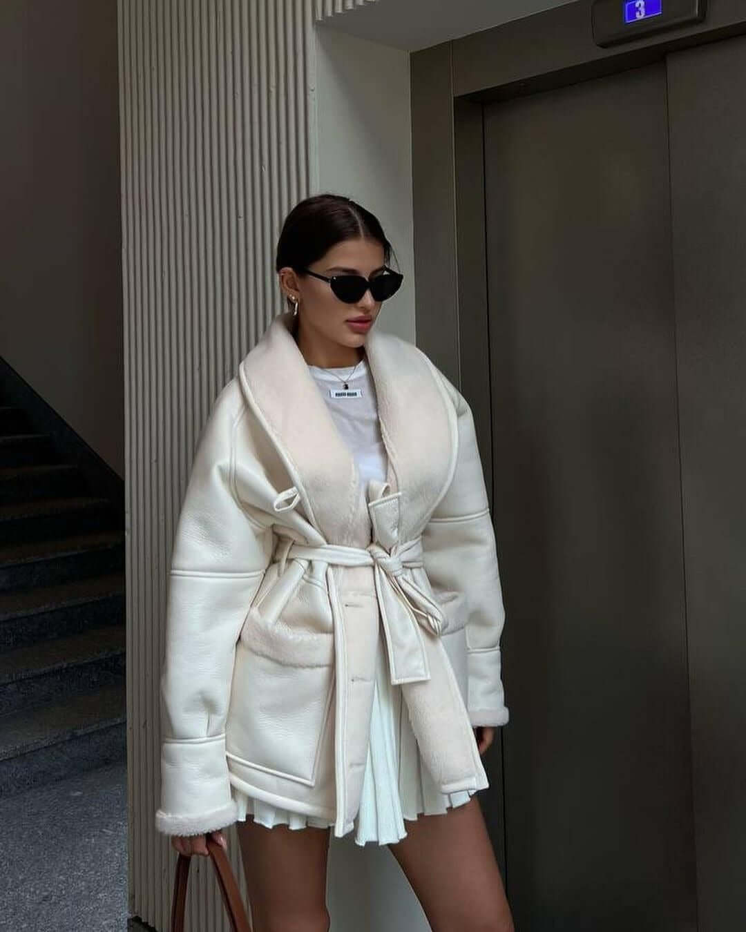 Woman wearing a stylish cream long sleeve coat with a belt, paired with a white skirt and sunglasses.