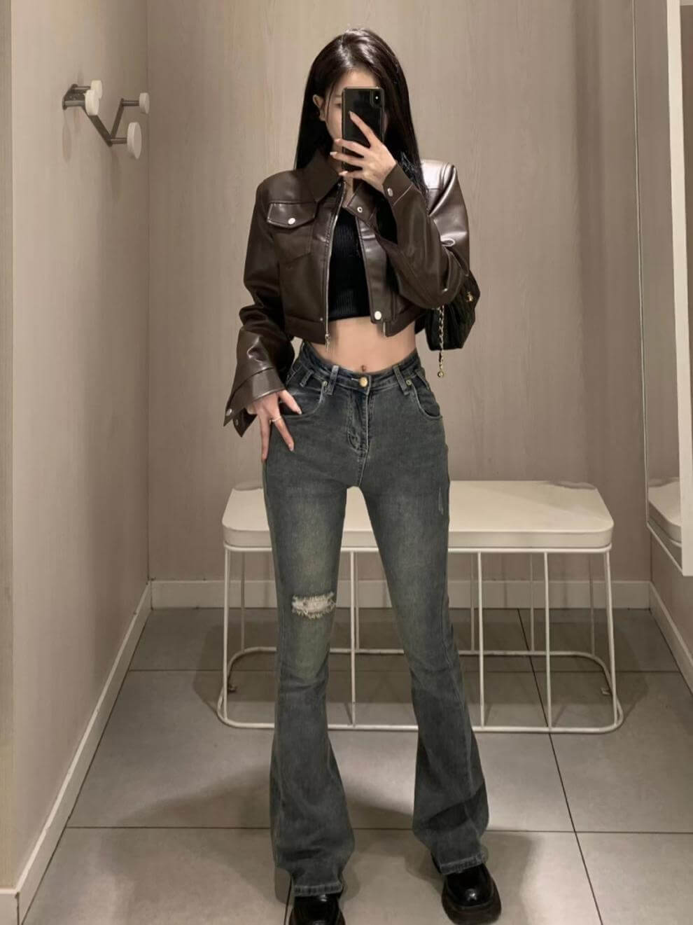 Stylish female wearing a coffee color leather motorcycle jacket with flared jeans in a modern setting.