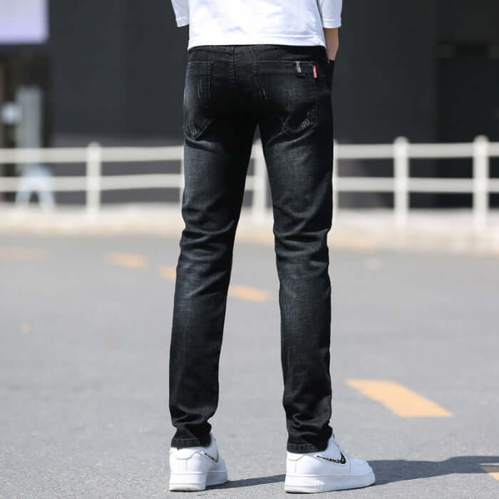 Men's slim straight black denim trousers viewed from the back, styled with a casual white shirt and sneakers.